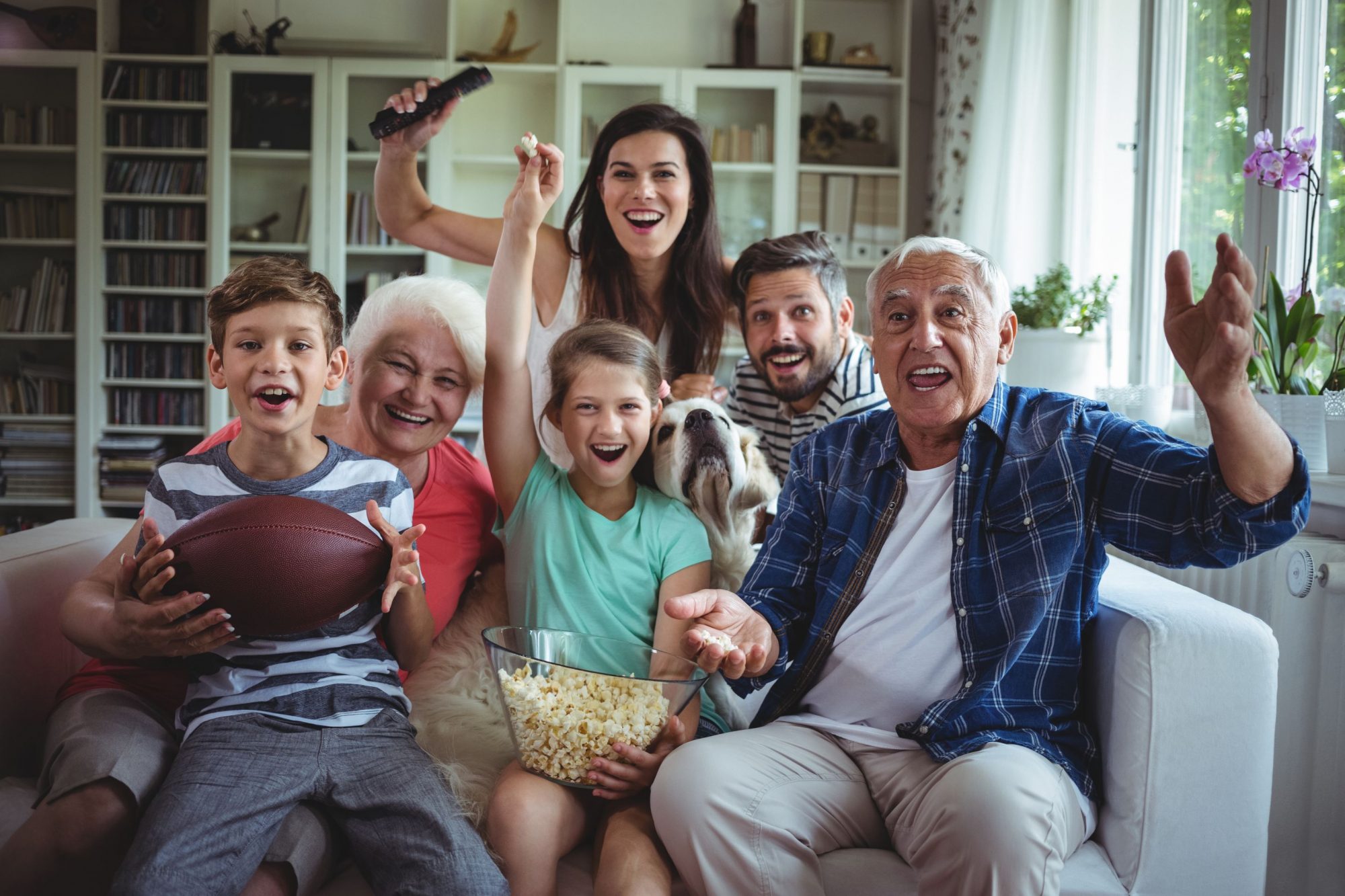 People on couch watching football with snacks and dog.